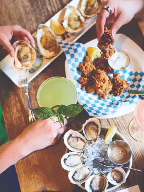 A table in Pioneer Square is filled with plates of oysters, fried seafood, dipping sauces, lemon wedges, and two cocktails. Two people reach for food—perfect for sharing and celebrating a World Cup match together.
