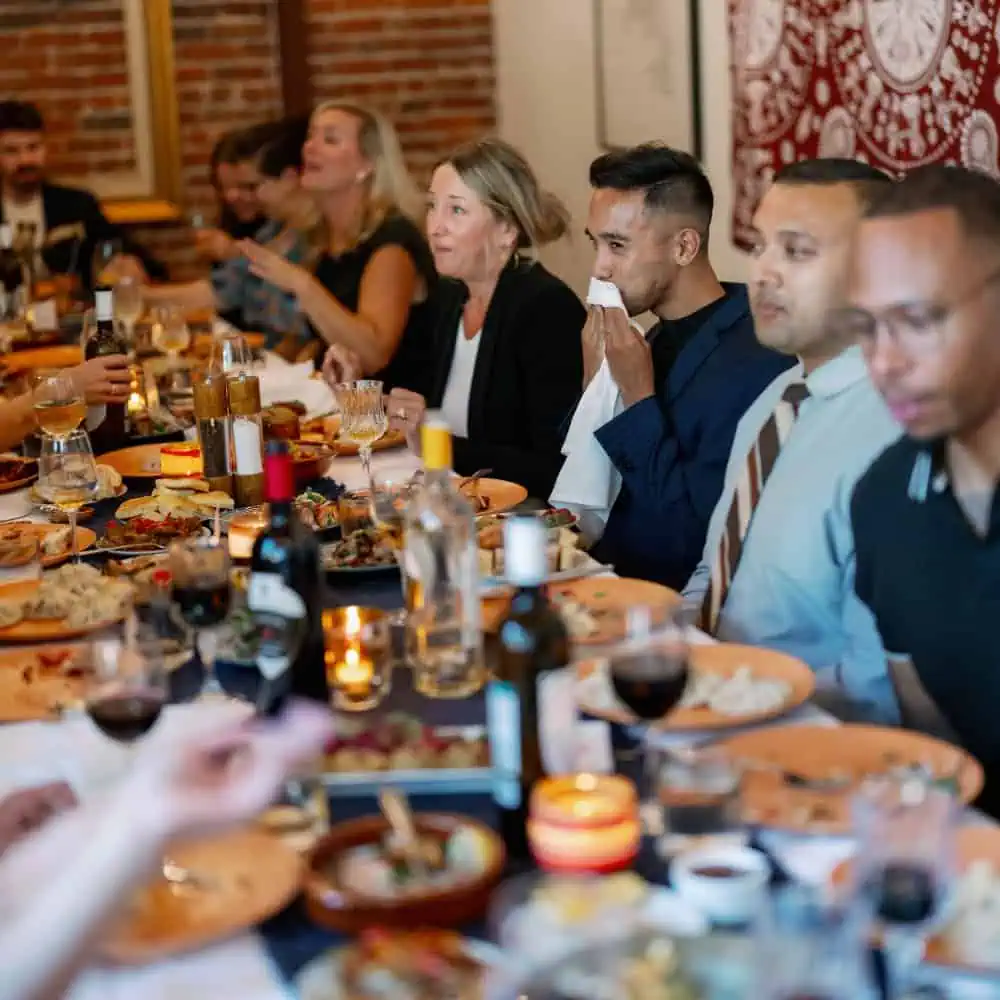 A group of people sit around a long table sharing a meal, with plates of food, drinks, and candles visible on the table.