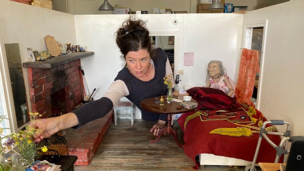 A woman arranges flowers inside a miniature room, with a lifelike elderly doll sitting in a bed next to a walker and a brick fireplace.