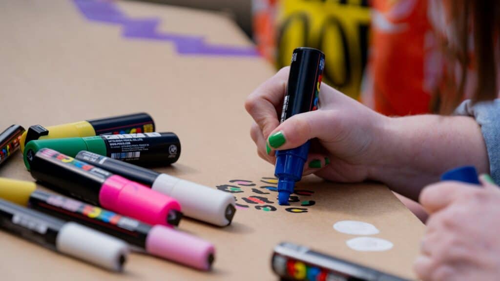 A person with green nail polish uses a blue marker to draw colorful shapes on brown paper, surrounded by various markers.