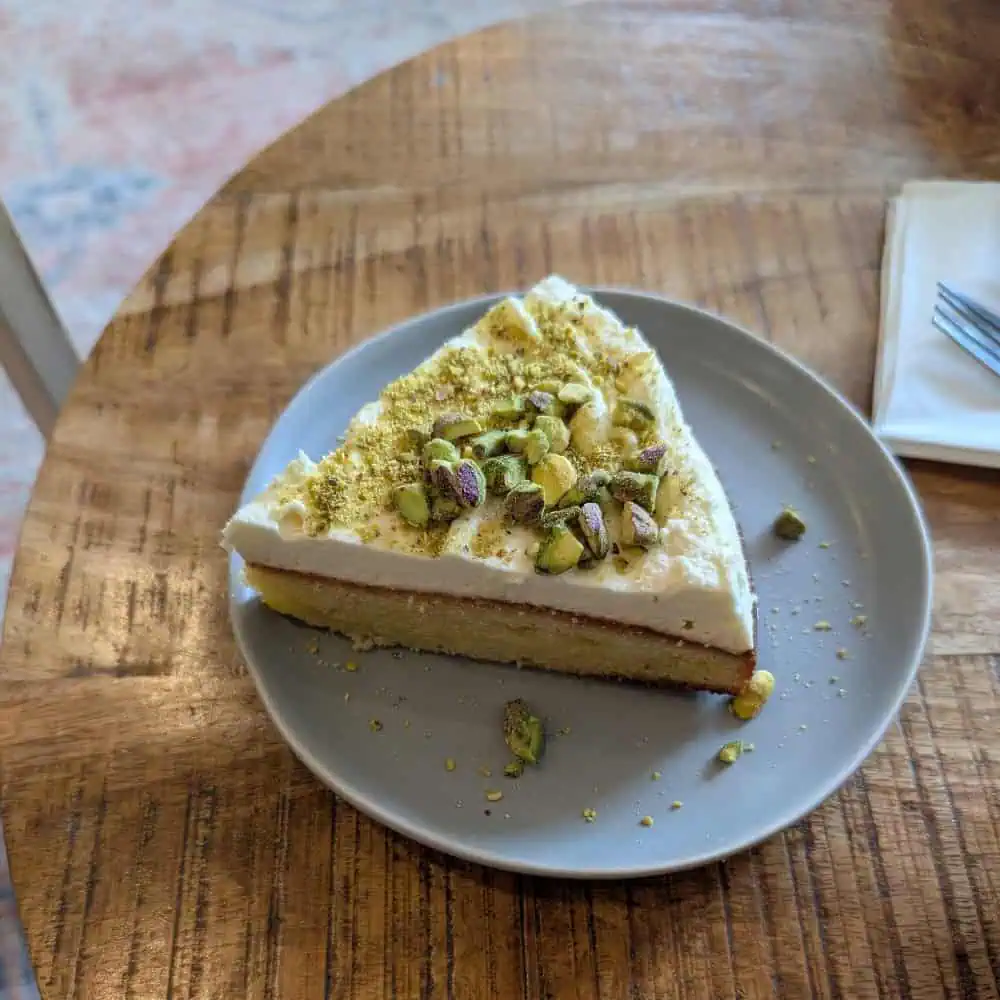 A slice of cake with white frosting and chopped pistachios on top is served on a gray plate on a wooden table.