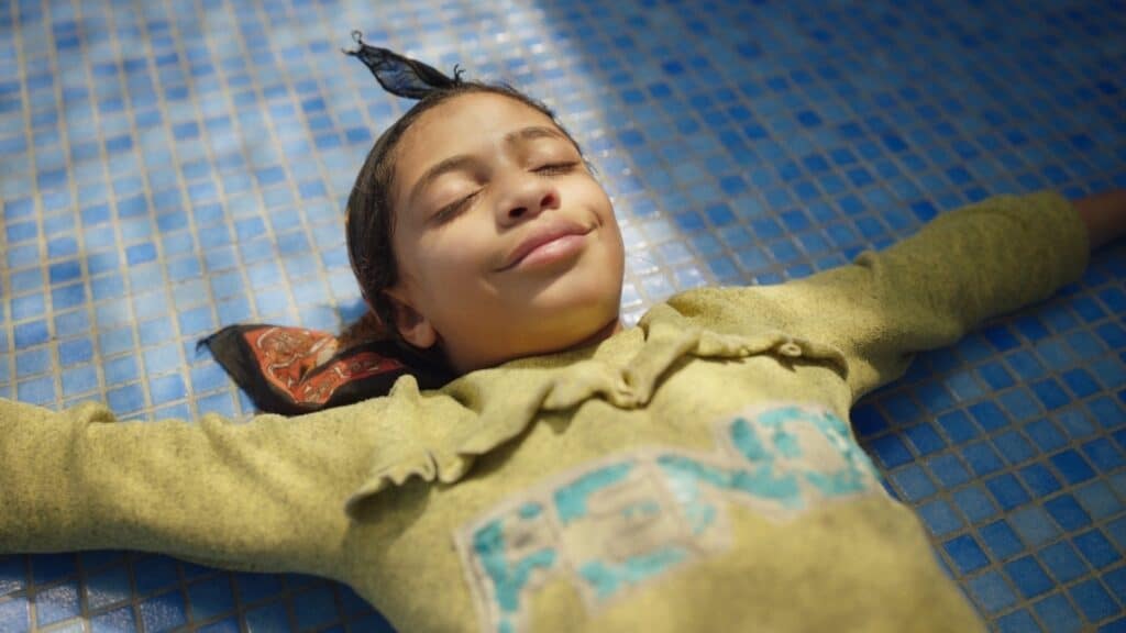 A child in a yellow sweater lies on blue tiles with arms outstretched, eyes closed, and a relaxed expression.