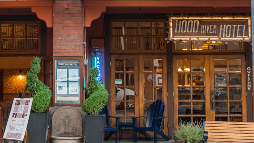 Front entrance of Hood River Hotel with a lit sign, glass doors, outdoor seating, potted plants, and information boards by the doorway.