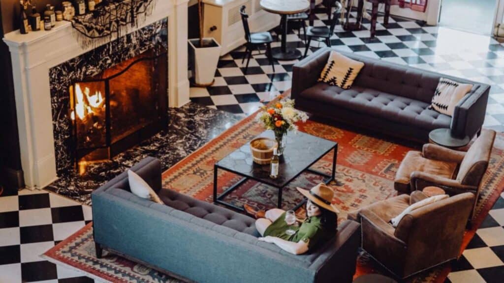 A person sits on a blue sofa in a cozy living room with a fireplace, checkered floor, area rug, and coffee table with flowers and a bottle.