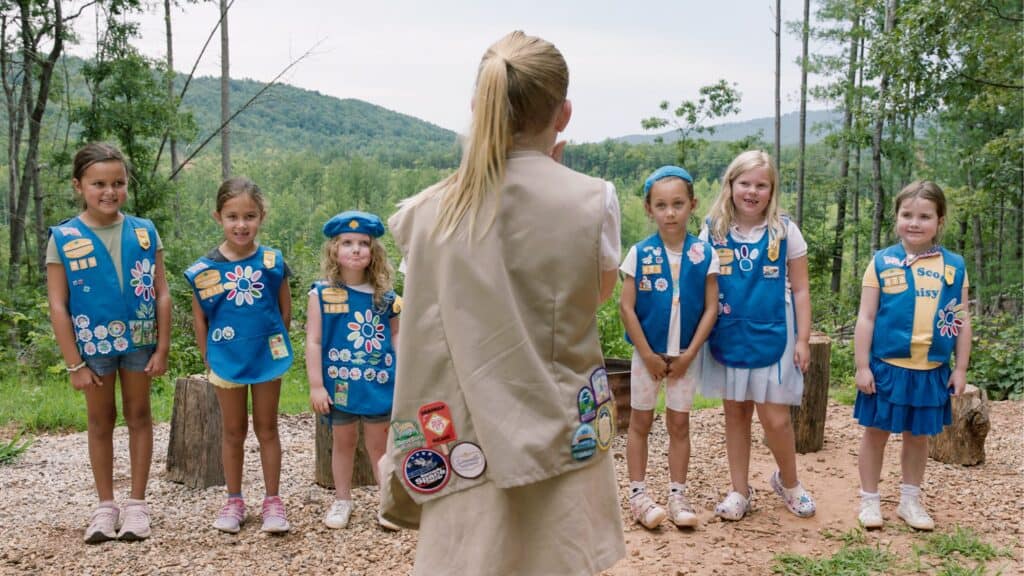 A group of young girls in blue vests with badges stand in a line outdoors facing an adult leader with her back to the camera.