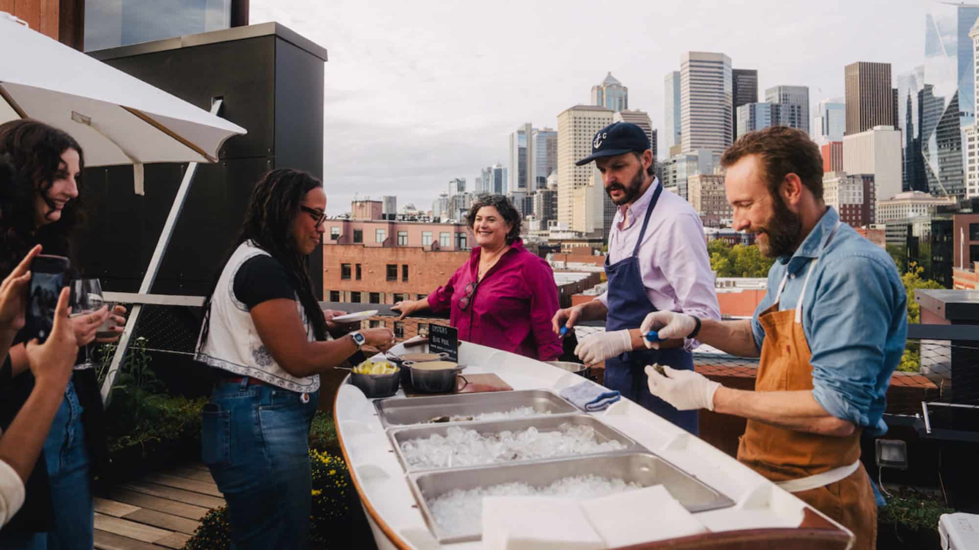 People gather at a rooftop bar where two staff members prepare and serve food from an ice-filled counter, with a city skyline in the background.