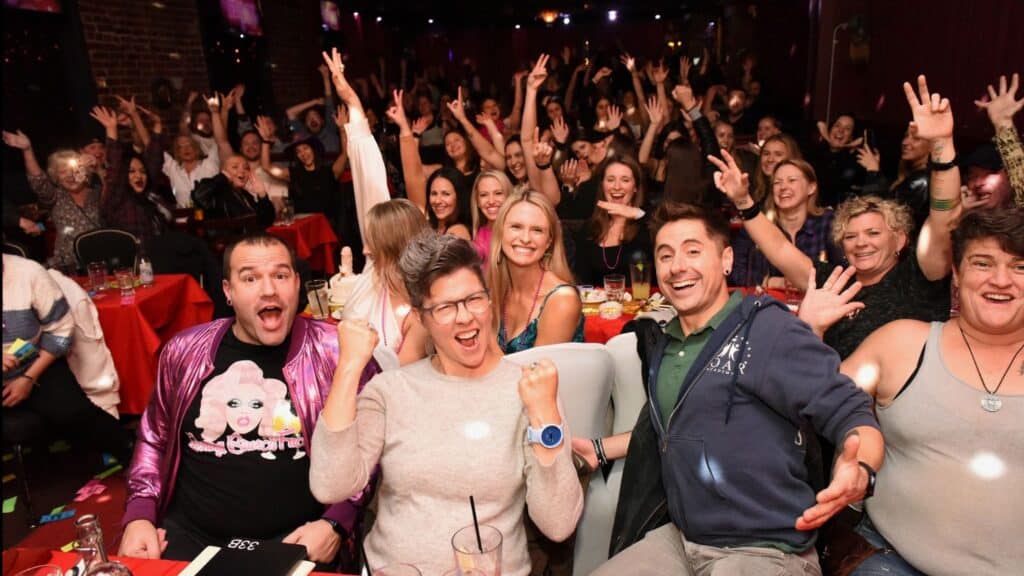 A large, cheerful crowd sits at tables in a dimly lit venue, posing with raised arms and smiling faces as they look toward the camera at a lively Seattle Drag Brunch.