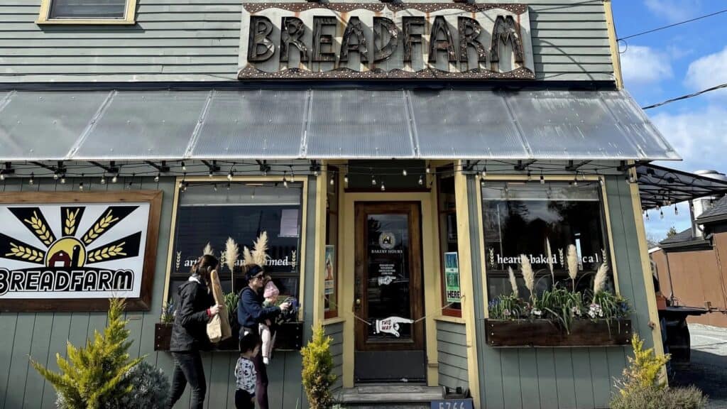 Exterior of Breadfarm bakery with two people and a child standing outside, plants by the entrance, and a large Breadfarm sign above the door—just a short drive from scenic Bellingham Bay.
