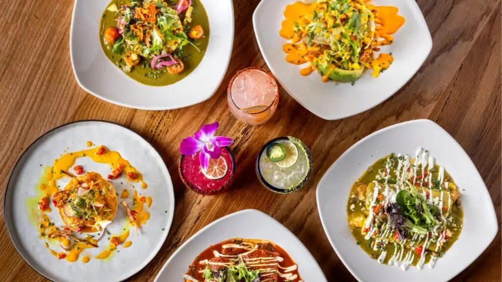 Overhead view of a table with multiple plates of Mexican food, including enchiladas and salads, and three colorful cocktails.