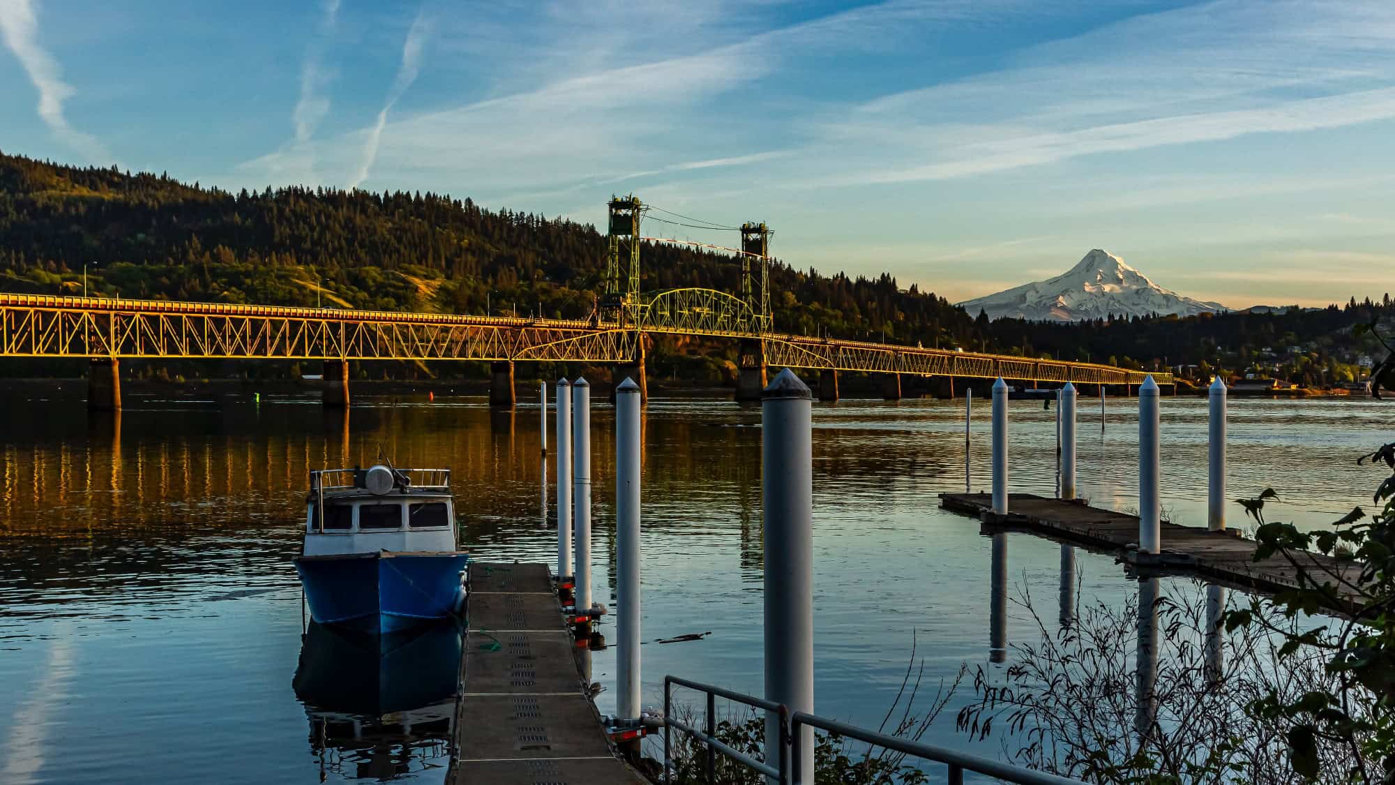 A boat docked on a river with a long bridge in the background, forested hills, and a snow-capped mountain under a blue sky with wispy clouds.