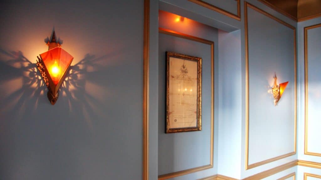 Wall-mounted sconces emit warm light onto light blue walls with gold trim; a framed document is hung in a recessed section of the wall.
