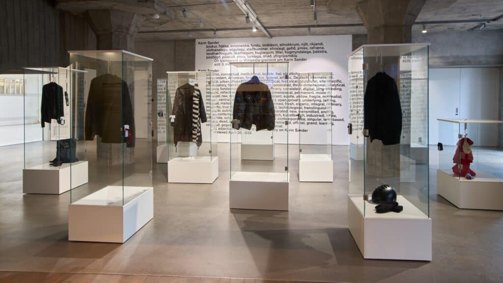 A museum exhibit displays jackets, shirts, helmets, and gloves in glass cases, with descriptive text on the wall behind them in a modern, industrial-style space.