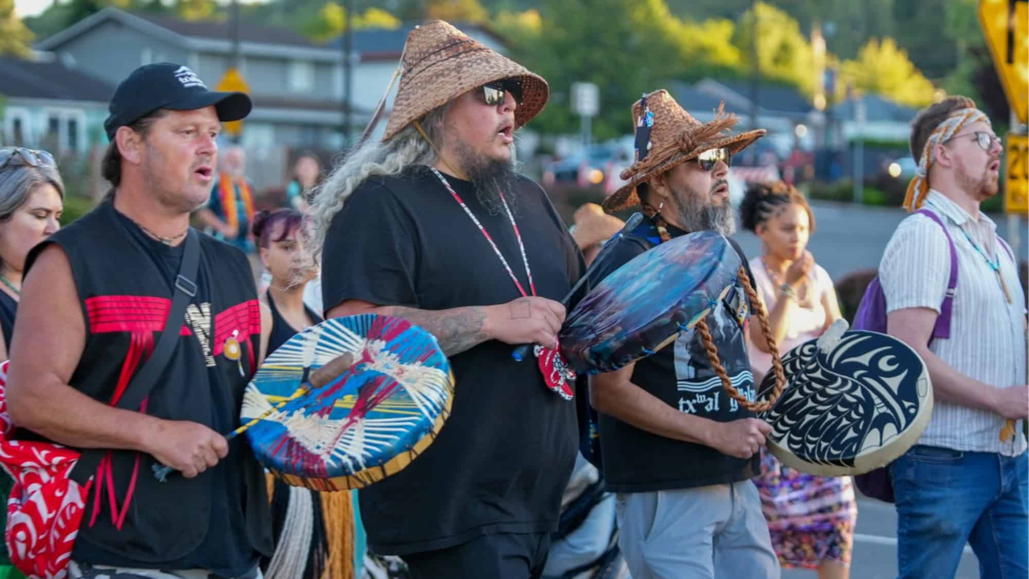 A group of men wearing woven hats and holding decorated drums walk together outdoors, with other people and buildings in the background.