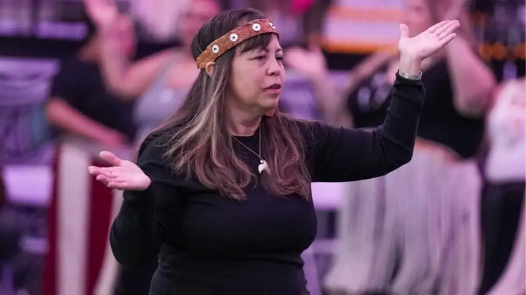 A woman wearing a brown headband and black shirt raises her hands while participating in an indoor group activity or event.