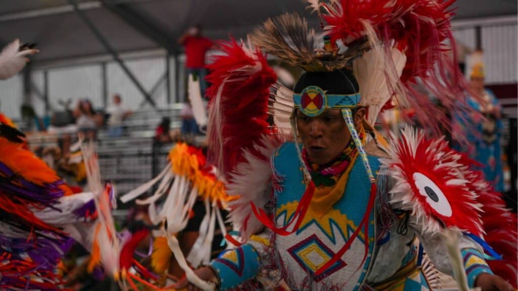 A person wearing colorful traditional regalia with feathers and geometric patterns participates in a dance at an indoor event.
