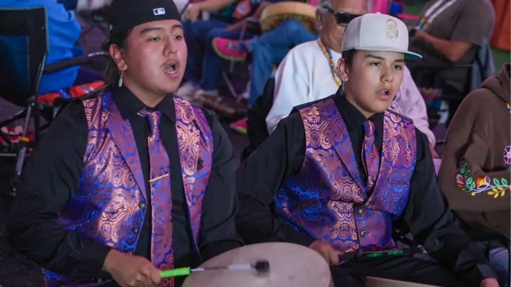 Two young people wearing patterned vests and baseball caps play a drum and sing at an indoor event.
