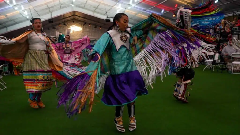 Several people wearing colorful traditional clothing perform a dance with fringed shawls inside a large indoor venue with a green floor and audience in the background.