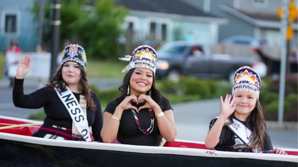 Three women wearing sashes and decorated headdresses, representing Coast Salish culture, ride in a Seattle parade float, smiling and waving at the camera.