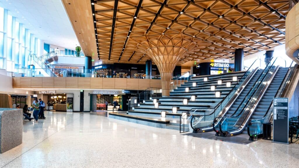 Modern airport interior with wood ceiling, wide steps, escalators, and shops including a café. Natural light enters through large windows on the left, offering striking airport views reminiscent of Icelandic Art.