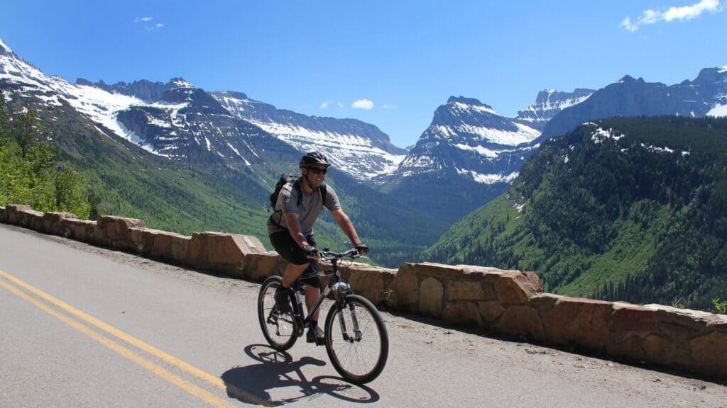 A person rides a bicycle on a paved mountain road with snow-capped peaks and green valleys in the background under a clear blue sky.