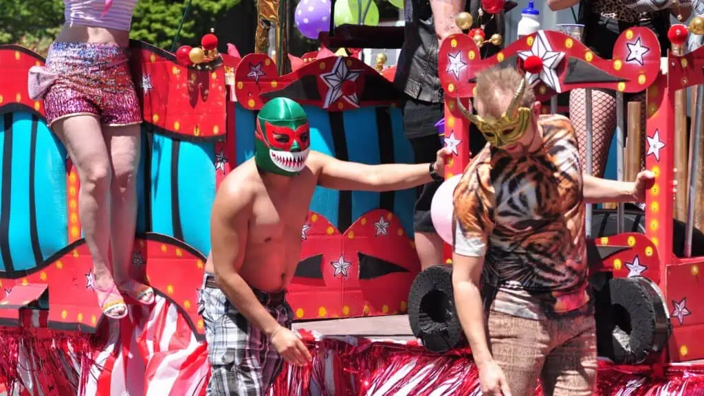 Three people on a colorful parade float; two wear lucha libre masks and one is shirtless. The float is decorated with red, blue, and star patterns.