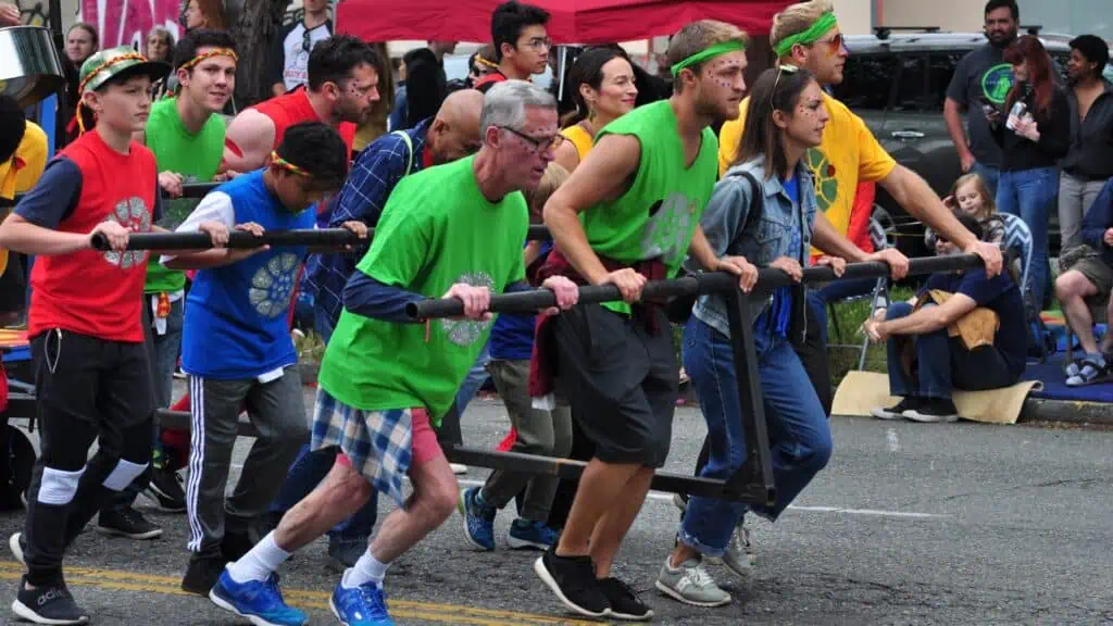 A group of people in colorful shirts participate in a team race, pushing a wheeled structure down a street, with spectators watching.