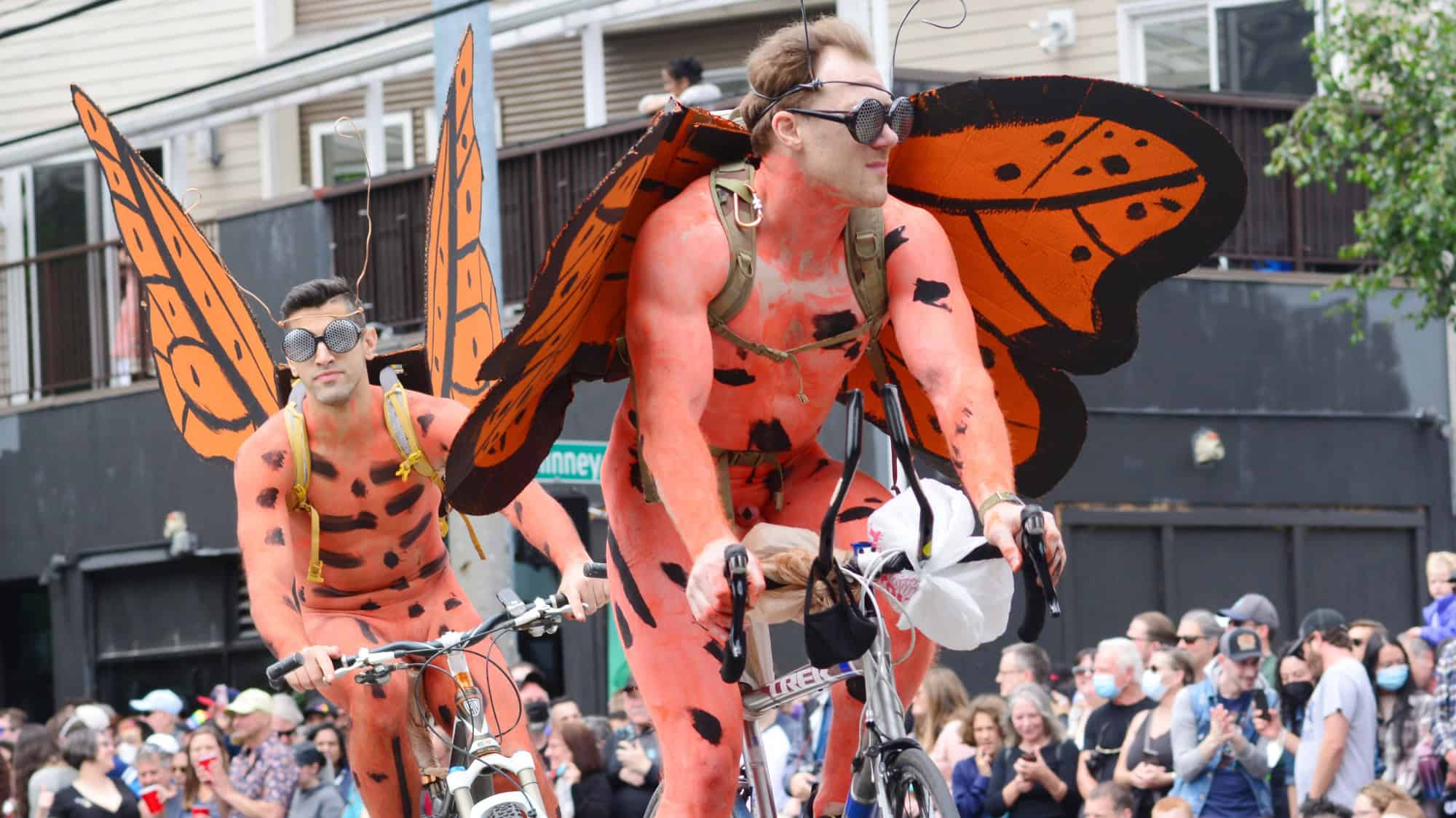 Two people in orange butterfly costumes ride bicycles in front of a crowd at an outdoor event.
