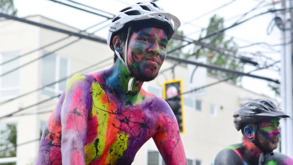 Two cyclists with helmets and colorful body paint participate in an outdoor event, with buildings and traffic lights in the background.