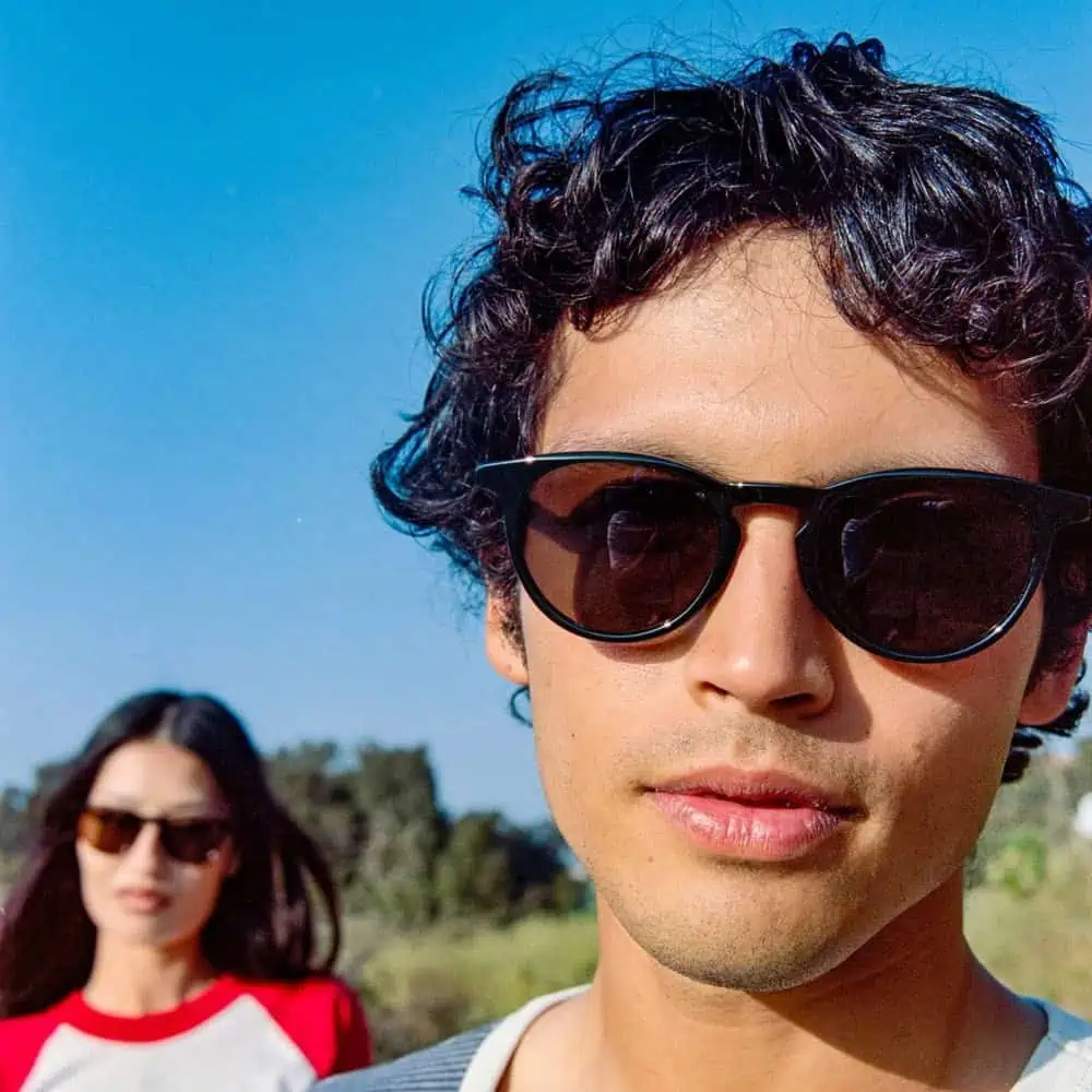 A man wearing sunglasses stands in the foreground, with a woman in sunglasses and a red-and-white shirt blurred in the background outdoors.