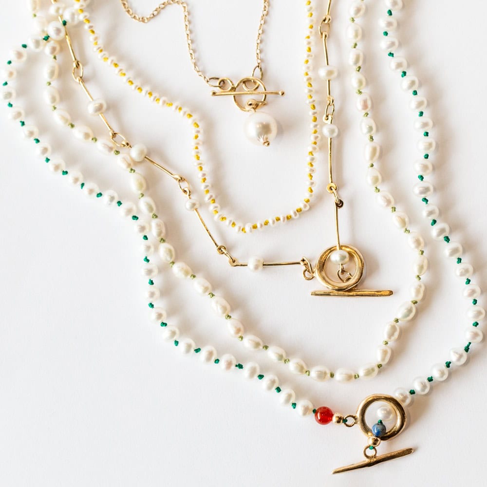 Four layered gold and pearl necklaces with different toggle clasps and bead accents are arranged on a white surface.