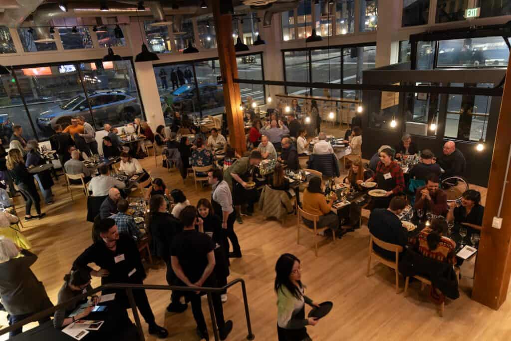 A busy restaurant with many people dining at tables, chatting, and walking around; large windows reveal a nighttime street scene outside.