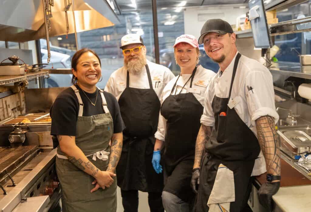 Four kitchen staff members wearing aprons and chef uniforms smile and stand together in a commercial kitchen.