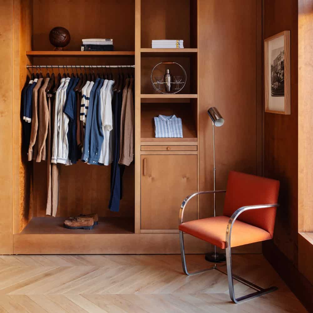 A wooden closet with shirts hanging, folded clothes on shelves, a football, a lamp, an orange chair, and a framed picture on the wall.