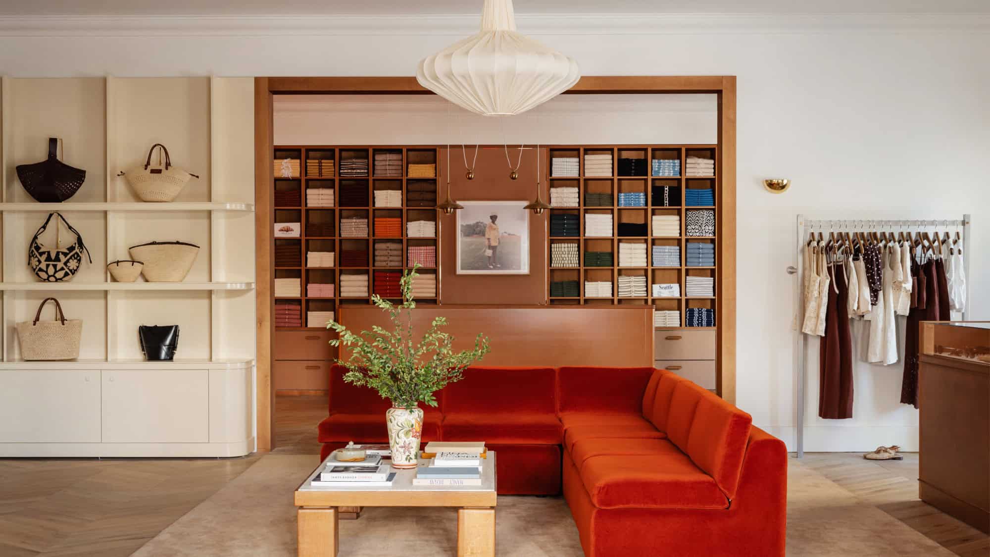 A modern boutique interior featuring a red velvet sectional sofa, a wooden coffee table with books, shelves with bags, clothing racks, and neatly arranged boxes in the background.