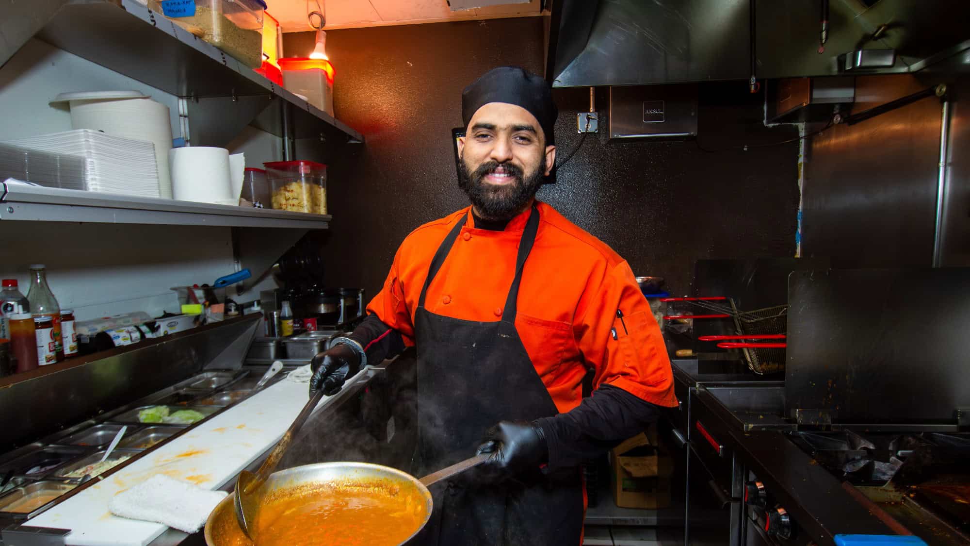 A chef in an orange uniform and black apron stirs a pan of sauce in a commercial kitchen, smiling at the camera.