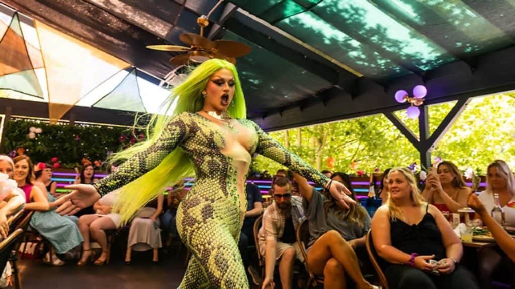 A drag performer in a green snakeskin bodysuit and neon green wig walks confidently past seated audience members at an indoor event.