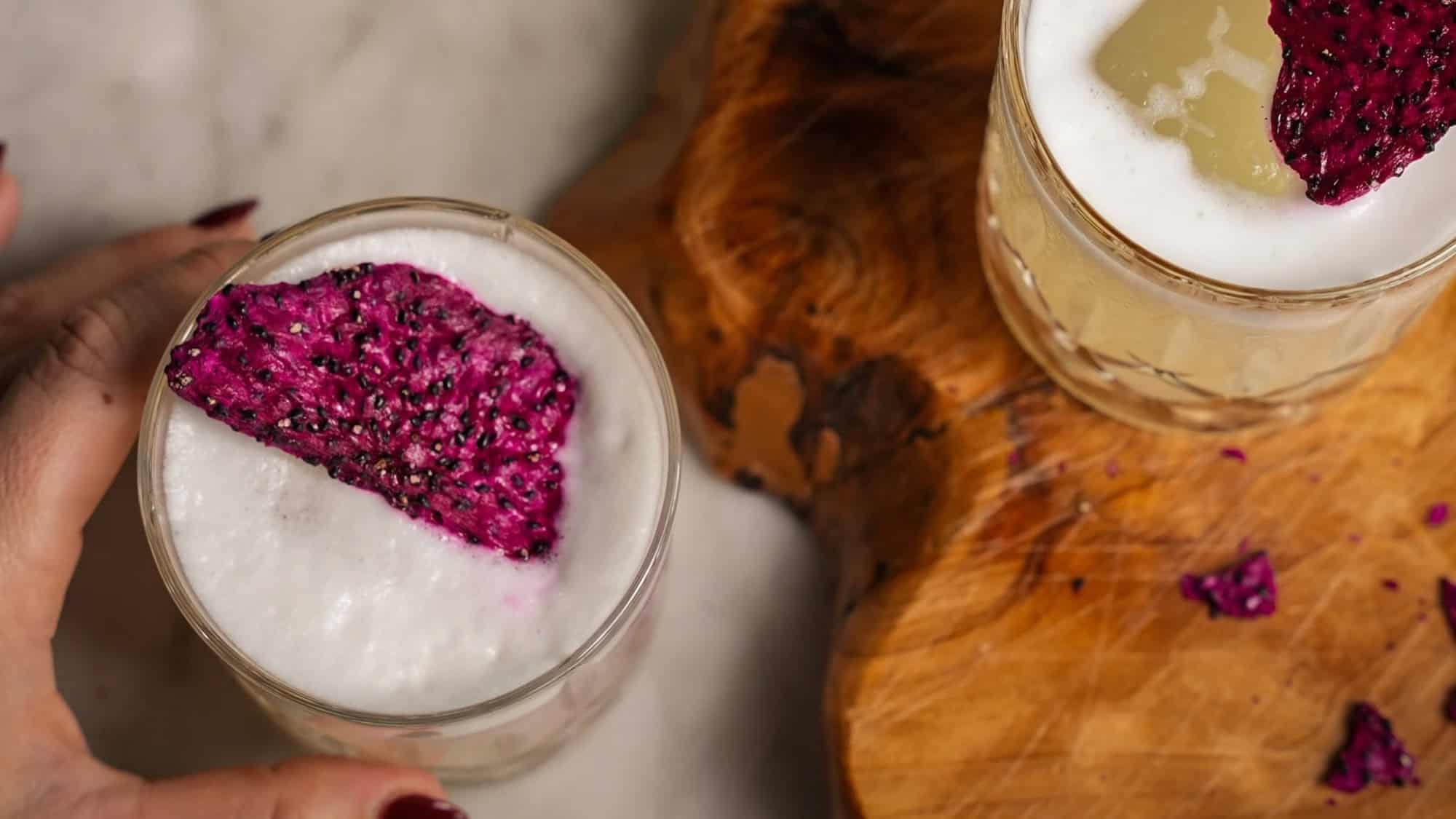 Two glasses with frothy drinks, each topped with a slice of dragon fruit, placed on a marble surface next to a wooden board.