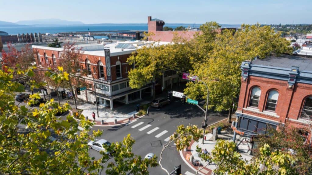 Aerial view of a downtown intersection with historic brick buildings, leafy trees, cars on the street, and a body of water visible in the background.