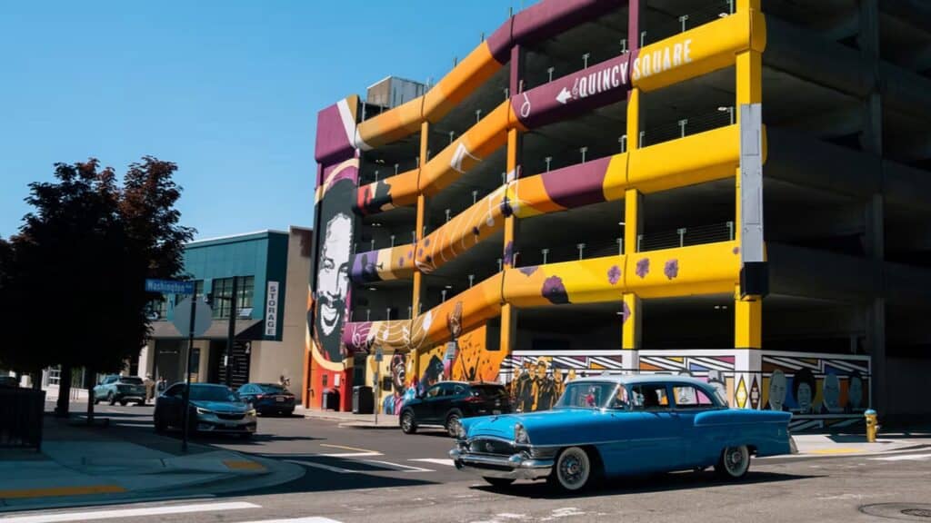 A bright yellow and purple parking garage with a mural is next to a road where a vintage blue car and other vehicles are parked or driving.