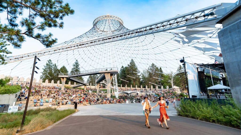 Two people walk in front of a large outdoor amphitheater with a crowd seated under a netted canopy structure, surrounded by trees and greenery.