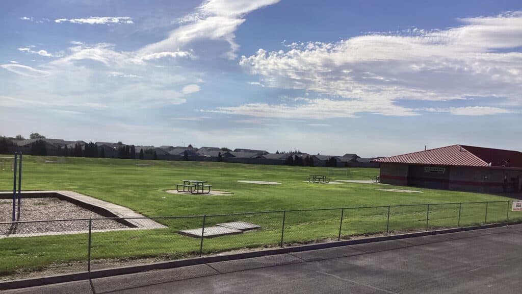 A large grassy field with picnic tables, a playground area, and a building with a red roof, bordered by a fence under a partly cloudy sky.