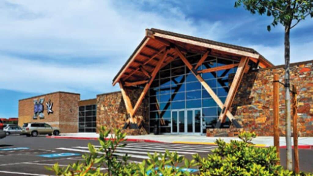 A modern building with large glass windows, stone walls, and a wooden entrance, seen from a parking lot under a partly cloudy sky.