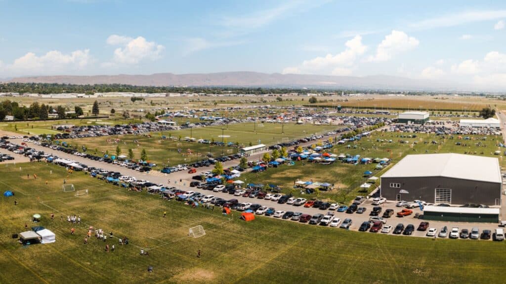 Aerial view of a large sports complex with multiple soccer fields, parked cars, tents, and groups of people attending an outdoor event under a partly cloudy sky.