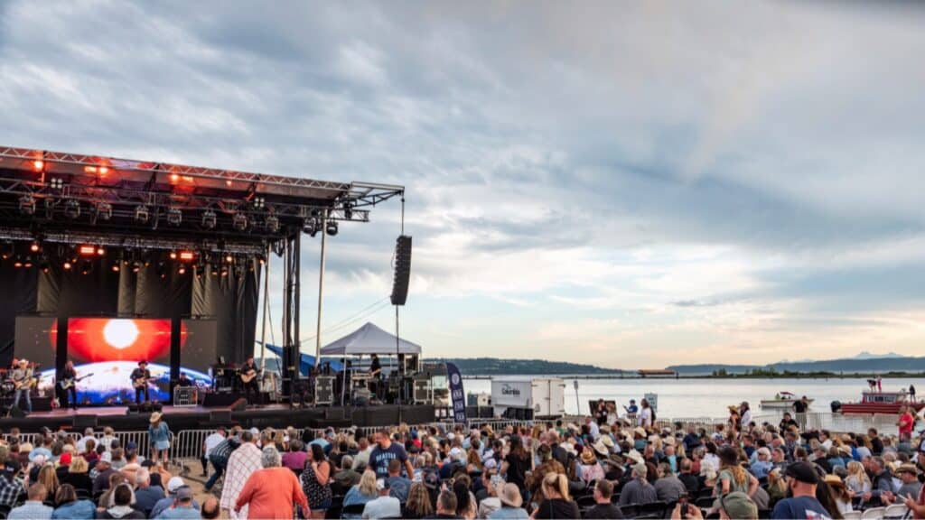 A large crowd watches a live outdoor concert near a body of water, with a stage, musicians, and a video screen under a partly cloudy sky.
