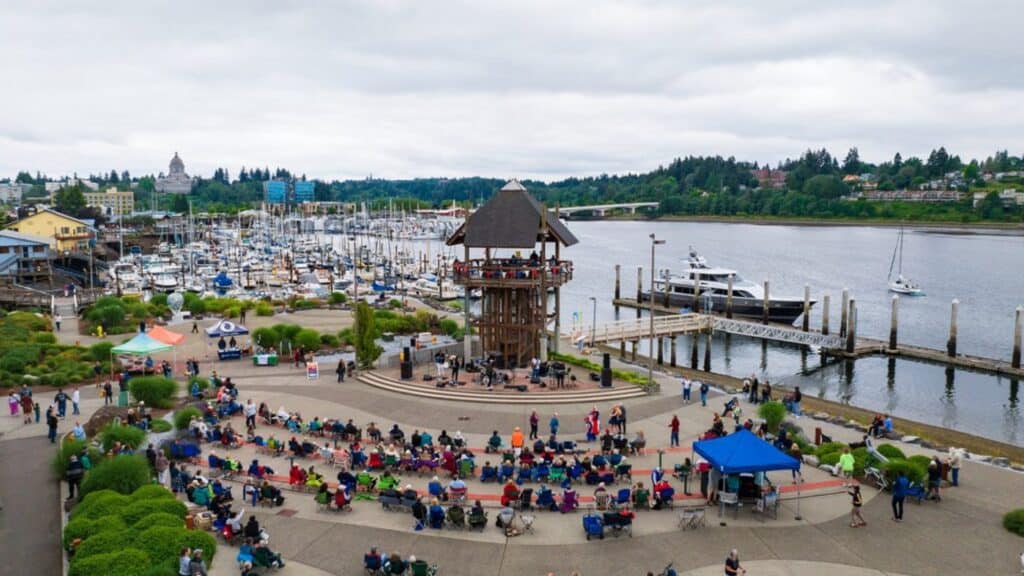 A crowd sits in front of an outdoor stage near a marina with boats docked along the water, under a cloudy sky.