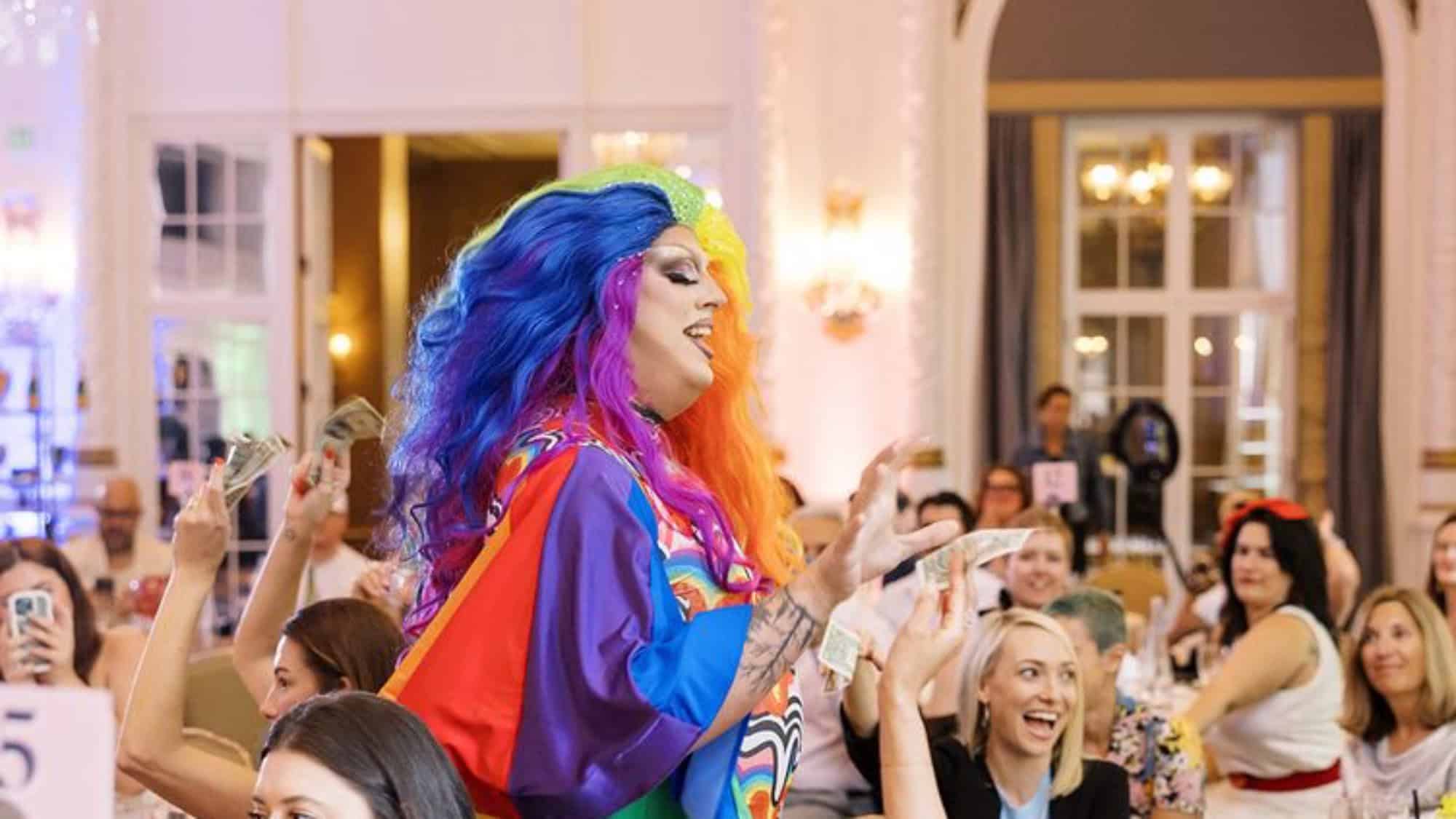 A drag performer in a colorful wig and cape interacts with a seated audience, some of whom are holding up dollar bills, at an indoor event.