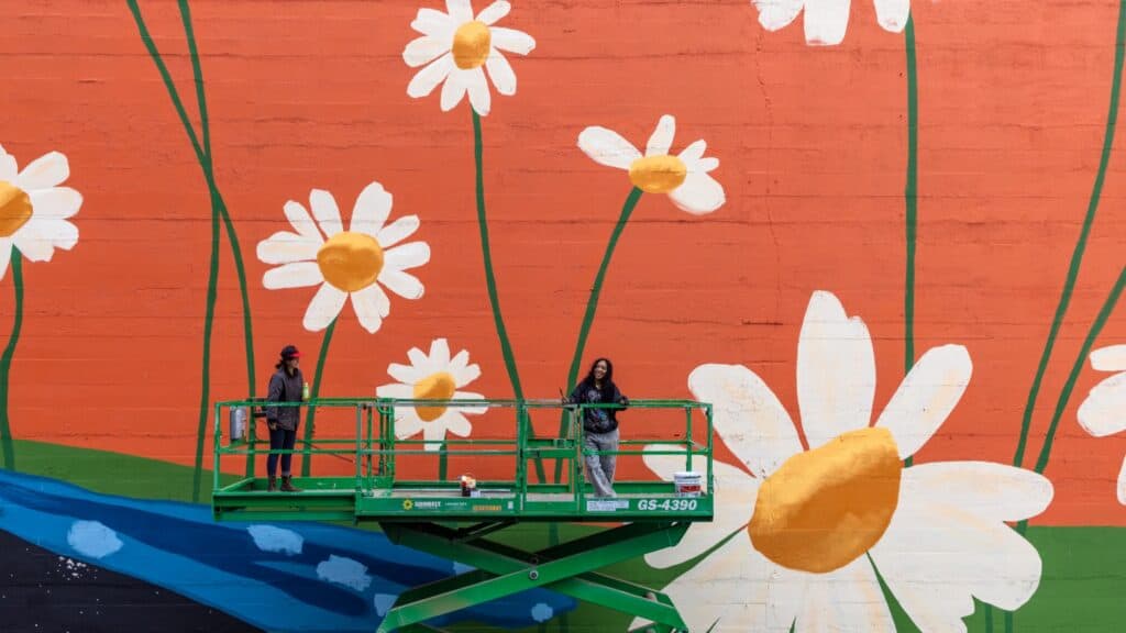 Two people stand on a green lift painting large white daisies with yellow centers on an orange wall.