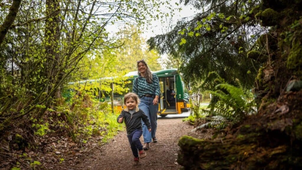 A child runs along a forest path near a Trailhead Direct stop, with an adult following behind, while a green and yellow bus is parked in the background.