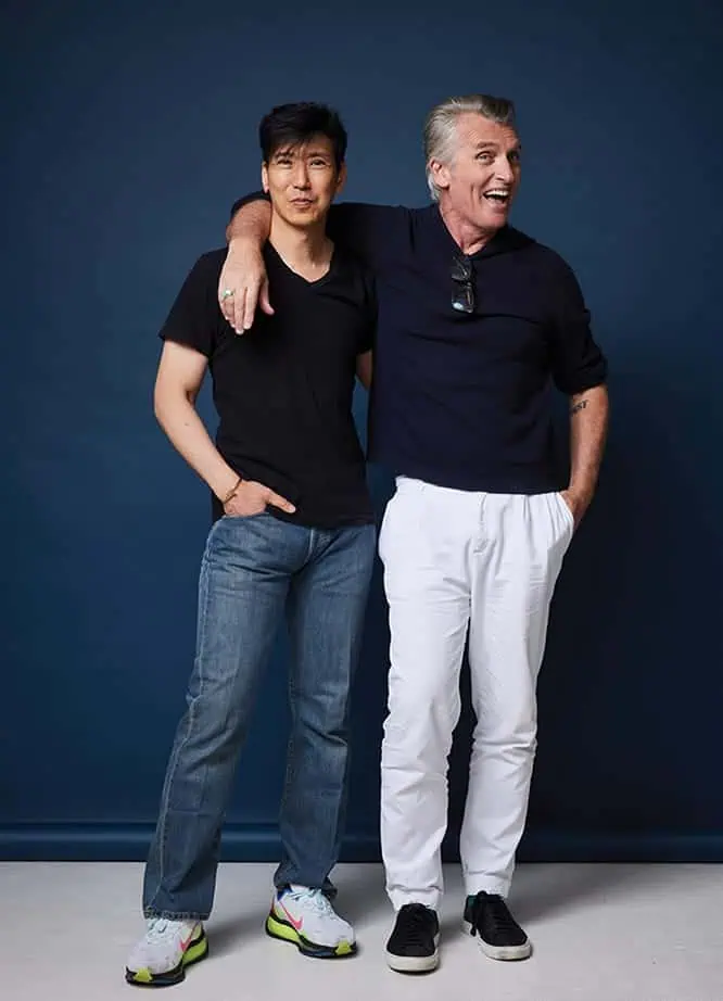 Two men stand together in front of a blue backdrop; one, possibly John Pearson, has his arm around the other. Their relaxed style in t-shirts, jeans, and sneakers reflects the easygoing side of fashion and culture.
