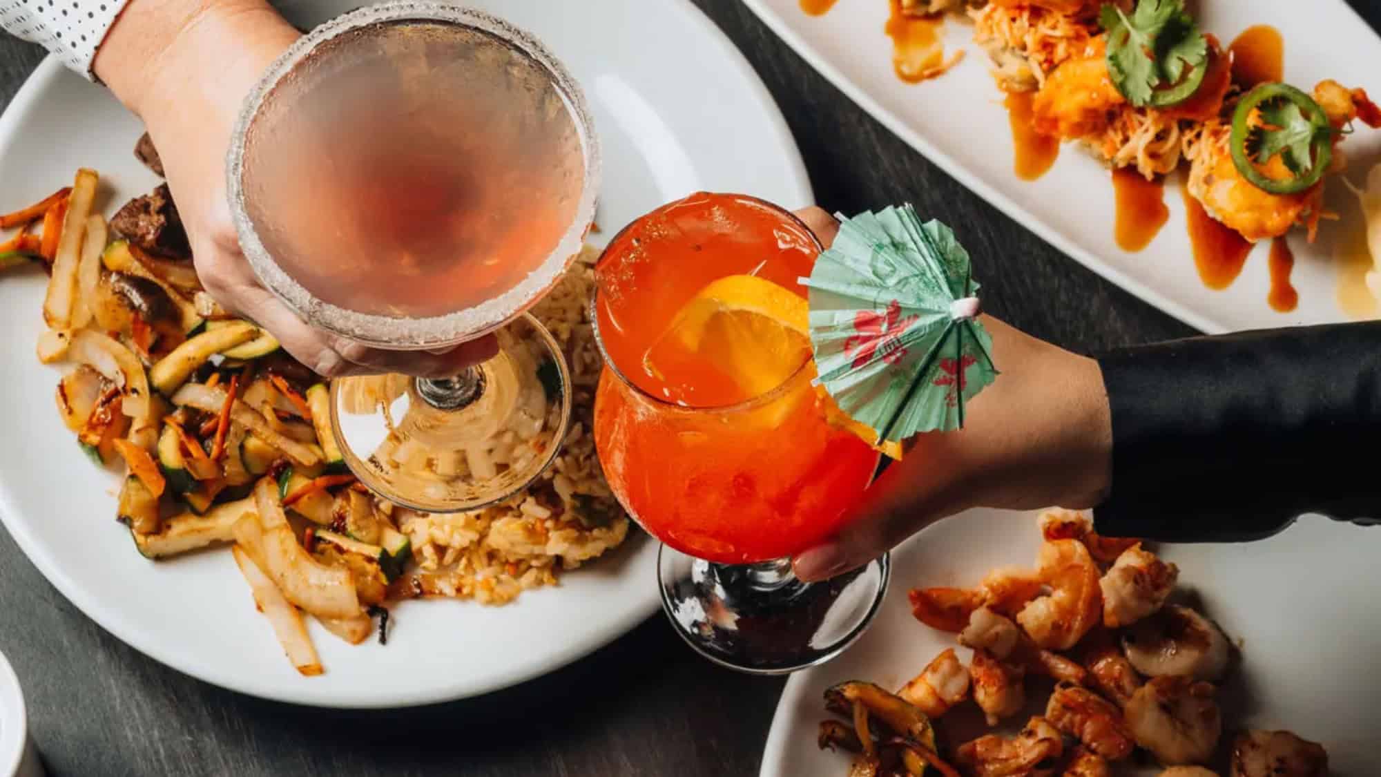 Two people clink cocktails over plates of assorted food, including stir-fried vegetables, shrimp, and Fantastic Frites, on a dark table at Benihana.
